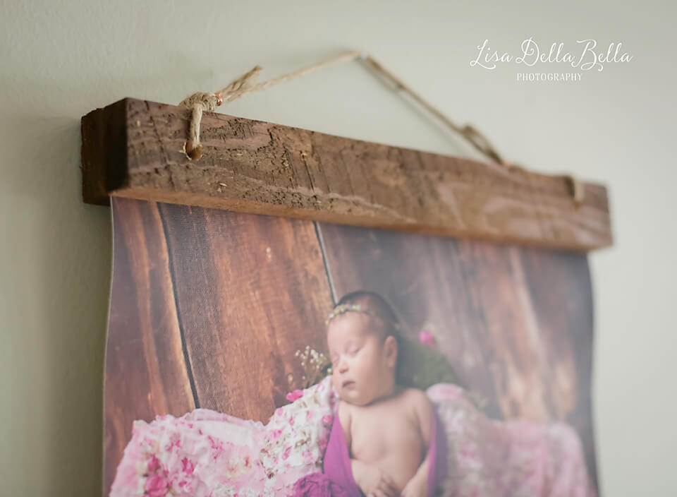 A close-up view of a photograph hanging on a wall, featuring a sleeping baby wrapped in a pink and purple blanket. The photo is secured with a rustic wooden frame and suspended by a rope, highlighting the delicate products within. The background includes a wooden texture. The watermark reads "Lisa Della Bella Photography.
